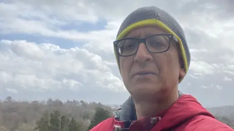 Dr Ravi Kumar - wearing black glasses, a green beanie and red jacket - taking a selfie with trees stretching into the horizon behind him