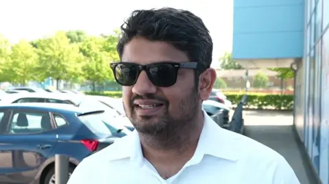 Arpit Gupta, has short black hair, dark sunglasses and a white shirt he stands outside the Manchester City Superstore.