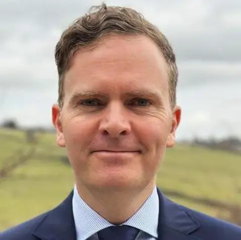 Reform UK A man with short brown hair wearing a blue suit and tie, standing in front of a field