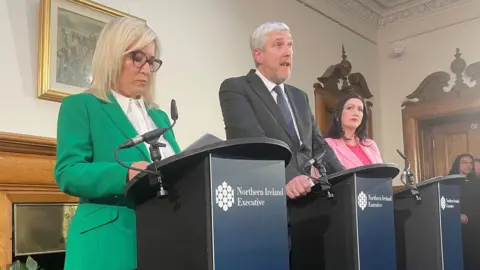 PA Infrastructure Minister John O'Dowd making the announcement on Wednesday with First Minister Michelle O'Neill (left) and Deputy First Minister Emma Little-Pengelly (right)