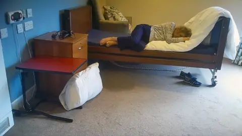 BBC A photo of an elderly woman on a bed in a care home room. The room is sparse with no personal effects. The floors are carpeted and one wall is blue. There is a bedside drawer and a portable desk. There is a pillow on the floor. Rachel lies on the bed in foetal position, fully-clothed, with her head on the white uncovered duvet.