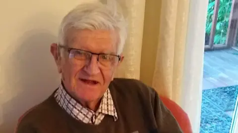 An old man with grey hair and glasses sits in a chair and smiles at the camera.
