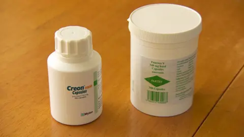 Two white tablet containers on a wooden table with labels reading "Creon Capsules" and "Pancrex". 