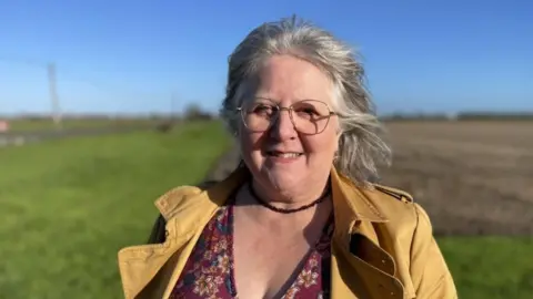 Lorna Dupre faces the camera and wears a yellow coat and a patterned flowery burgundy top. In the background is a blurred-out field.