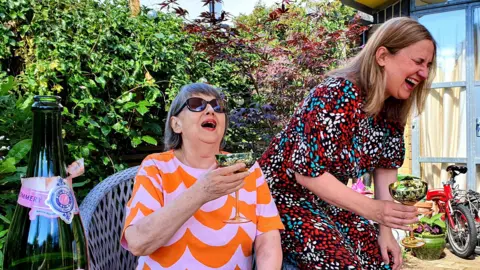 Handout A woman with short grey hair, an orange and purple print top and sunglasses sits in a garden chair and grins while holding a glass of champagne. Perched on the arm of the chair is another woman, with longer light brown hair and wearing a red, black and blue print dress, she is laughing and has her eyes closed.