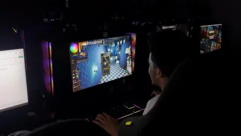 Kian Boyle/BBC A student is seen in a dark room sat at a computer along a bank of other computers playing a video game.