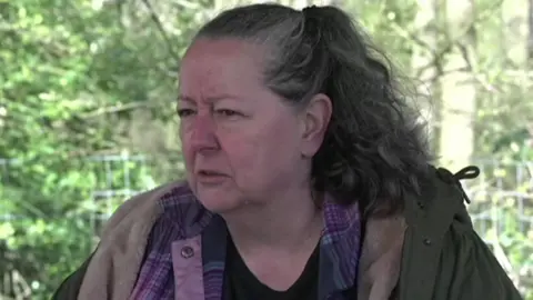 A woman with long dark hair and wearing a green coat over a purple flannel shirt over a dark t-shirt, looks to her right. Trees and greenery can be seen behind her.
