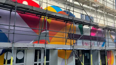 A building with three levels of scaffolding against it. There are three lots of double doors, with windows. On the door on the right hand side there is a sign which says 'UNITY' in red. There circles of paint on the wall. The colours include blue, orange, yellow, purple and red. 