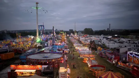 The Hoppings at dusk with rows of light up rides, the tallest being a tower with swings flying around the top. There are rows of white caravans to the right and a red Chinese noodles stall in the foreground.