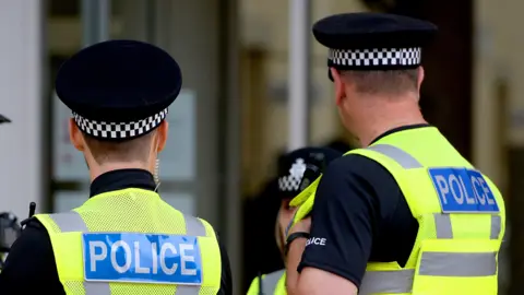 Two male police officers stand with their backs to the camera. On their high visibility vests, the word Police can be seen in white writing on a blue background. Both men wear peaked caps with a chequered band around them. 