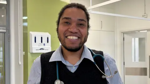A man with long brown curly hair smiling tied up smiling in the camera. She is wearing a shirt and jumper with a blue and white striped shirt with collars and has a stethoscope round his neck.
