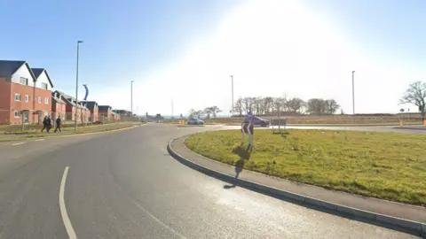 Google A roundabout, a tarmac road with some houses in the distance.