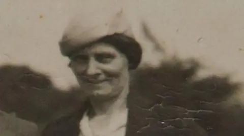 Louisa Dunne in a black and white photograph. She is wearing a hat and a brown jacket with a white shirt. 