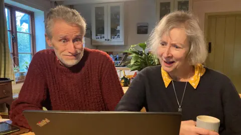 A man and woman, both with greying hair, are sitting at the kitchen table of their home, looking in frustration at a laptop. He is wearing a dark red jumper, she a navy blue one over a yellow blouse.