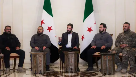 Sana Ahmed al-Sharaa wearing a dark suit and white shirt sitting in front of two Syrian flags with with three men in dark clothing and one in military uniform from Syria's new joint forces.