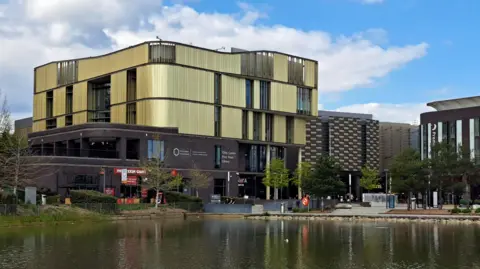 A four storey, gold cladded building, with a lake in the foreground. Trees and a footpath line the lake.