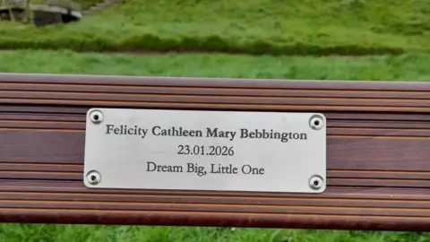 Jade Morris A park bench with a plaque on it. The text reads 'Felicity Cathleen Mary Bebbington. 23.01.2026. Dream Big, Little One'.