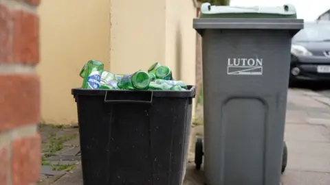 Qays Najm/BBC Two bins sitting on a pavement in a residential street. The bin on the left is a black plastic box, which is full of empty green bottles. To the right is a grey bin, with a green lid and "Luton" written on the front.