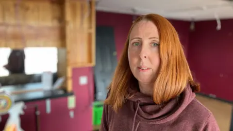BBC A woman in a red hooded sweatshirt stands inside an empty restaurant. It has no chairs and tables and the bar behind her is empty.  
