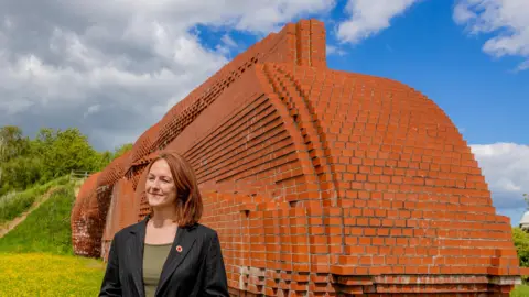 Lola McEvoy, Darlington MP, stands smiling next to the Darlington brick train. She is wearing a green top and a black suit jacket.