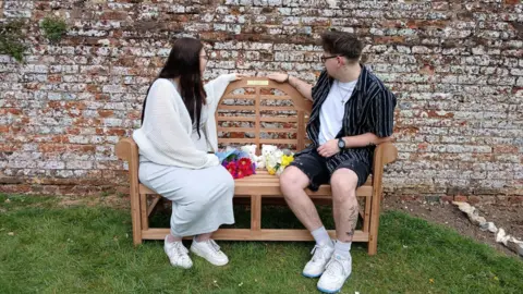 Hampshire Hospitals A man and a woman sat on a bench looking at a gold plaque which is on the top of the bench. Between the pair are two bunches of flowers, one red and one yellow with two teddies also between them.