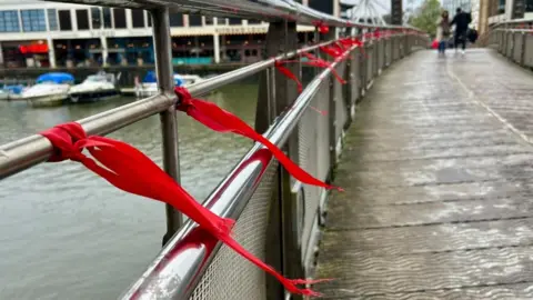 About a dozen red ribbons have been tied to the railings of a bridge, with boats visible on Bristol Harbour below.