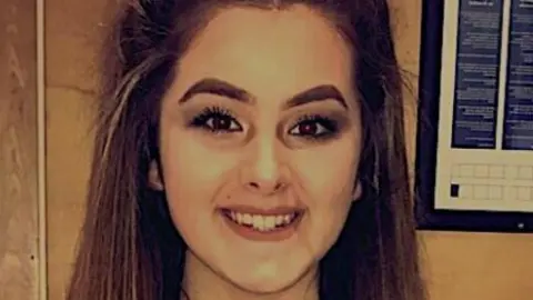 Emily smiles at the camera. She has dark eyeliner on and has thick long brown hair.