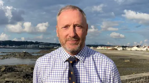 Paul Montague - a man with short grey hair and a short grey and brown beard looks at the camera. He is wearing a blue and white chequered shirt, with a dark blue tie with insignia's on it. In the background is a coastal view of houses and buildings.