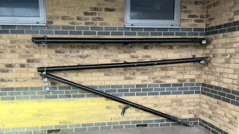 Barnwood AFC Three black poles beneath two windows on a brick wall at a football club, which are meant to be horizontal. However, one end of the pole on the bottom is touching the tarmac below, and the one above it is also skewed.