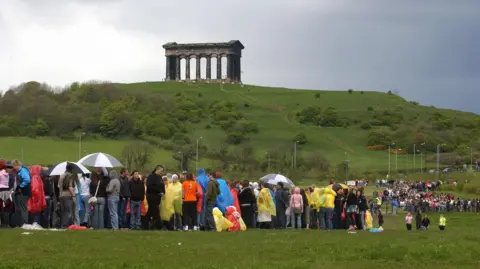 A long queue of people in lots of different coloured raincoats and holding several umbrellas. They are standing in a field with hills in the background. At the top of the hill is the Penshaw Monument which was built in the style of an ancient Greek temple. 