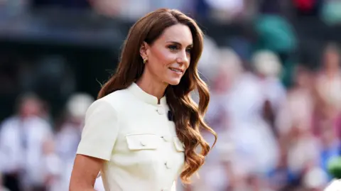 PA Media The Princess of Wales is in centre of the image, with her hair worn in long brown curls and wearing a thick gold hoop earring. She is wearing a very structured cream button-up top, with short sleeves and a high neck.  
