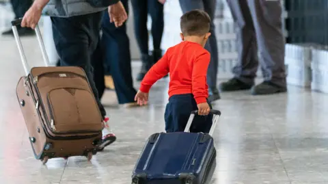 A young boy wearing a red jumper pulling a blue suitcase through an airport. There is an adult pulling a brown case walking in front of him.