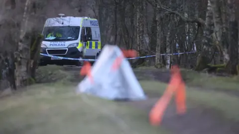 A police van is parked on a path in a wooded area. A small tent covering evidence is in the foreground.