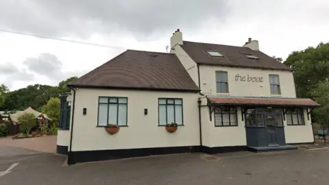 A cream two-storey building with the boat written on the front. The building is seen from the roadside at the front. It has window boxes with flowers and blinds pulled down. A carpark is behind it, with a tented garden area just visible.