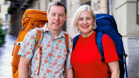 A man and a woman standing next to each other. The man is wearing a short-sleeved shirt with lobsters on it while the woman is wearing a red T-shirt. They are both smiling and looking into the camera while wearing large rucksacks on their backs.