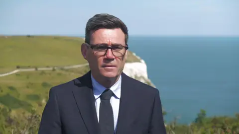 Mid shot of Andy Burnham wearing a black suit and black tie with black-framed glasses. In the distance is the white cliffs of Dover.