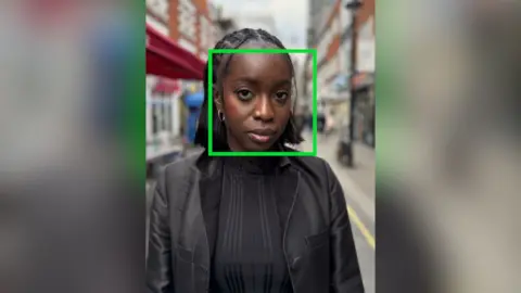 A woman in a black jacket stands in the street. A green box outline highlights her face