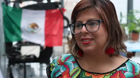 Erika Servin Gonzalez is speaking to an interviewer off camera. She has mid-length brown hair and is wearing glasses and red tassel earrings. Her top is multi-coloured and there is a Mexican flag hung up behind her.