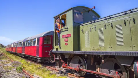 Alderney Railway Society A diesel train engine pulling two London Underground carriages on a grassy track with a path to one side. A smiling older woman leans out the cockpit in a navy train driver's uniform and high visibility orange jacket.
