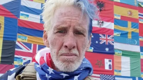 Reuters man stands shooting a selfie with blue and grey hair, a military vest and an American flag neck scarf. A large pattern of flags are behind him as a backdrop