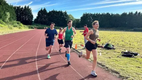 Drew Mclellan Four people running down the inner lane of the red Whitchurch track. They are wearing sports clothes including vests and shorts.