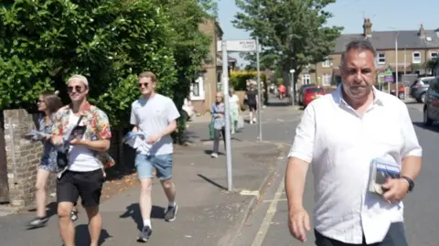 Steve Tuckwell with canvassers walking on the pavement to the left of him on sunny day in residential street. He is holding leaflets with his left hand and is walking on the road.