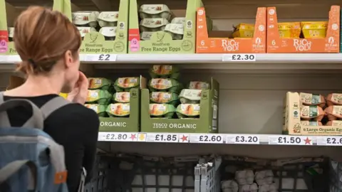 A woman with tied-up brown hair is facing away from the camera looking at a supermarket shelf. She is wearing a long sleeved black top and has a blue rucksack on her back. On the shelves is several different coloured packets of eggs.