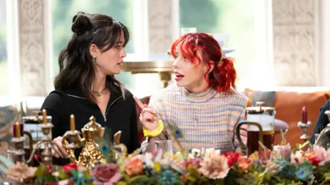 Studio Lambert/Euan Cherry/PA Two women captured mid-conversation as they sit in an elaborately decorated room with bright windows behind them. Jade, on the left, has shoulder-length brown hair, which has been gathered back into two space buns to either side of her head. She is wearing a black jumper. Jessie, on the right, is wearing a multi-coloured jumper. She has shoulder-length red hair which has been gathered into two bunches on either side of her head. They are sitting at a table with flowers in the foreground.