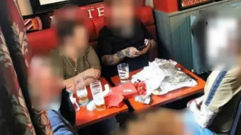 BM A group of five people, whose faces have been blurred, sit around a table at a pub. They have pints of beer in front of them and bags of crisps, as well as red scarves and other paraphernalia.