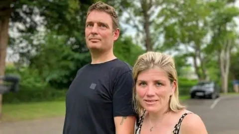 A man with brown hair and a black top stands next to a woman with blonde hair.