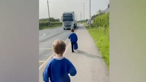 Two children in blue school jumpers walking down the road towards the primary school about a metre away from a lorry driving down a main road