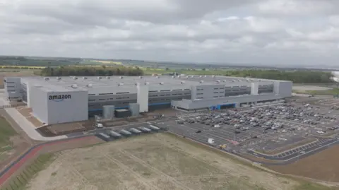 Amazon A sprawling grey box building, which is four storeys high, stands next to a car park. The building has an Amazon sign in the corner and is surrounded by fields.