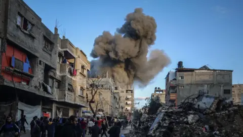 Smoke billows upwards in front of a blue sky and grey buildings. 