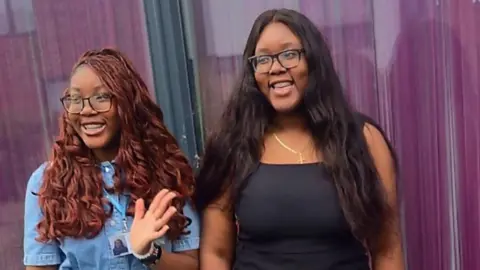 Tito Adeyale Two young women posing together outside a building, standing in front of large glass windows or doors. Tito on the left wears a blue denim shirt, and on the right Adeola wears a black top.